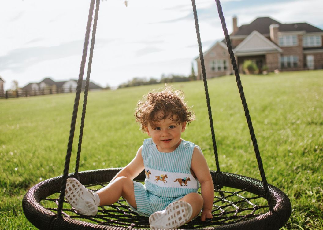 Charlie, a two year old boy with curly hair, sits on a swing outside his parent's house.