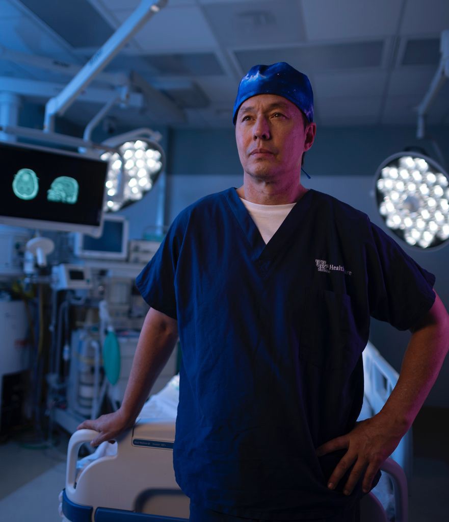 An Asian, male, doctor stands with hand on his hip in an operating room. A monitor with brain scans is in the background with other medical equipments and overhead lights.