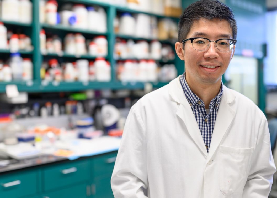 Ronnie stands in his lab coat in front of shelves packed with medicine bottles.